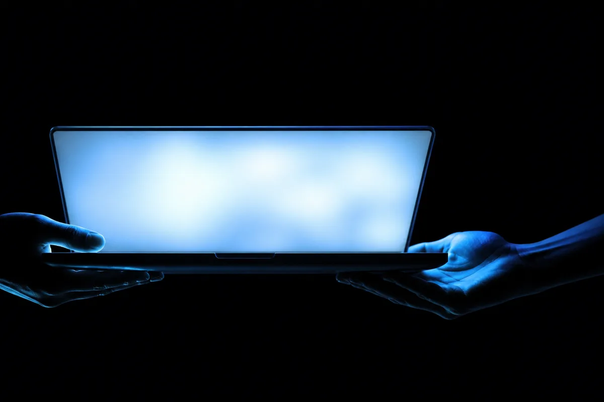 Dua orang di meja kerja, satu menyerahkan laptop terbuka kepada yang lain dengan kedua tangan